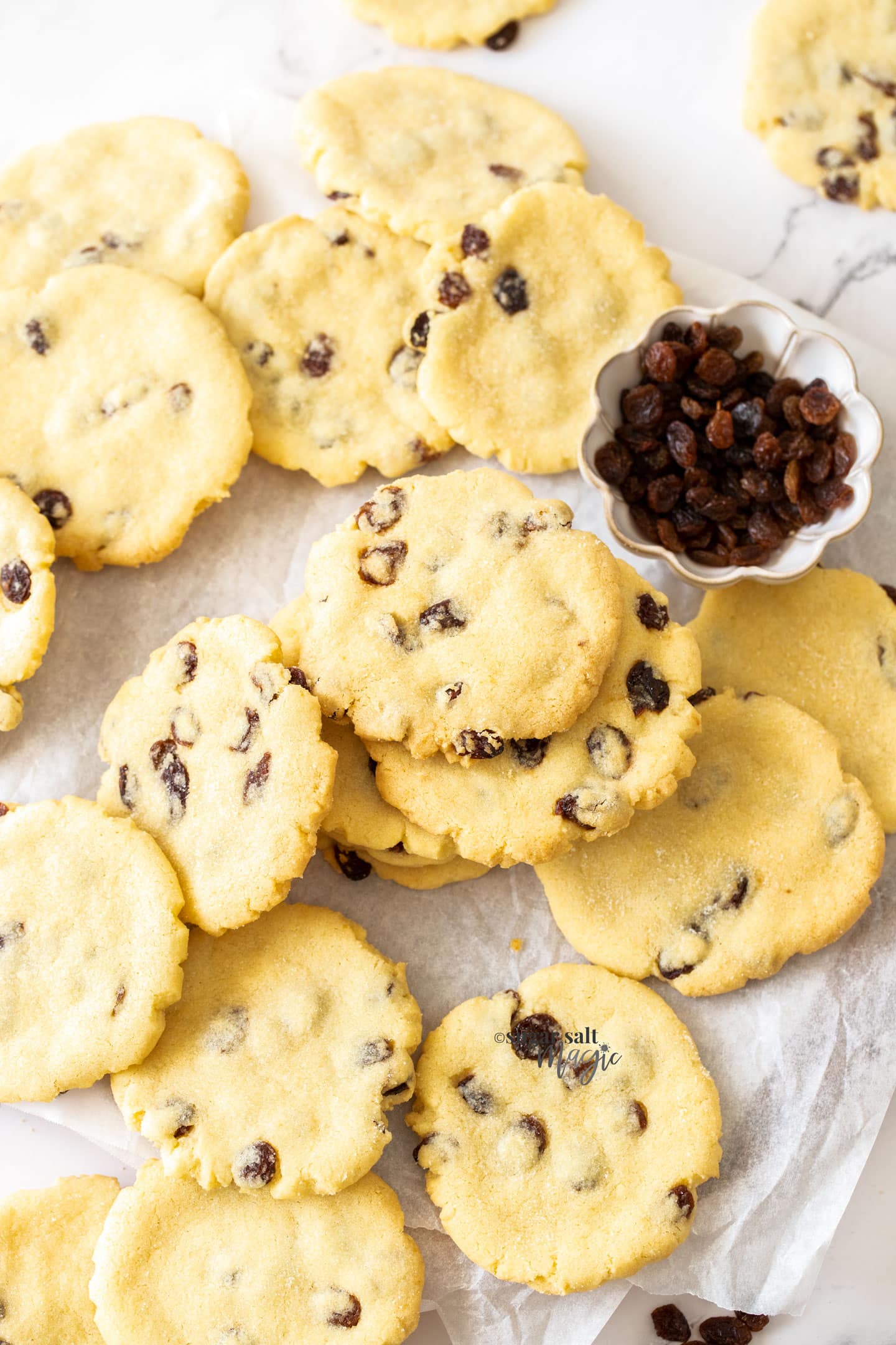A pile of sultana filled cookies with a small bowl of sultanas.