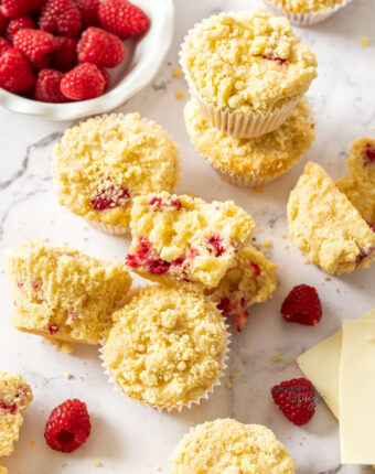 A batch of muffins with raspberries around them on a marble benchtop.