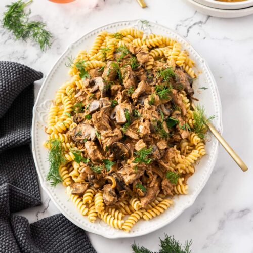 A white dish filled with pasta and beef stroganoff, with gold cutlery.