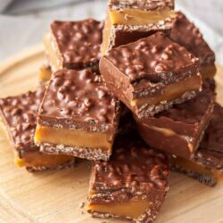 A pile of chocolate caramel slice on a wooden board.