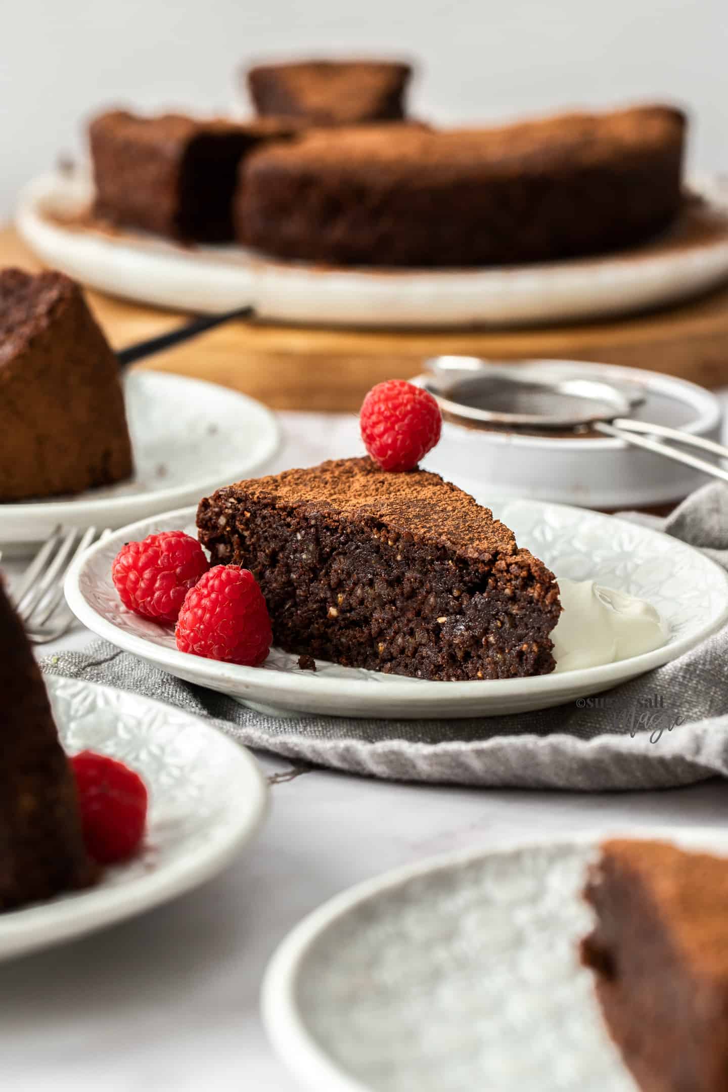 A slice of chocolate cake on a white cake plate with cream and raspberries.