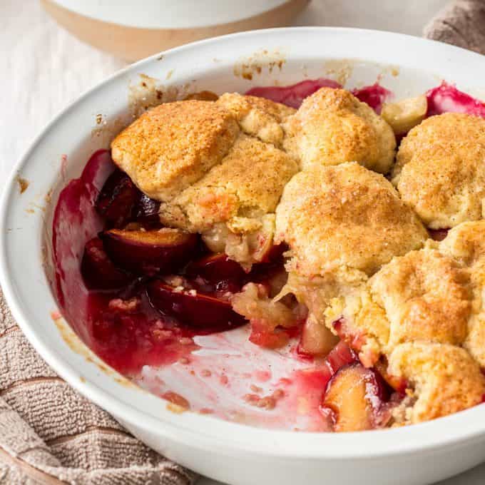 An apple and plum cobbler in a white pie dish with a scoop taken out showing the stewed fruits under the biscuit topping.