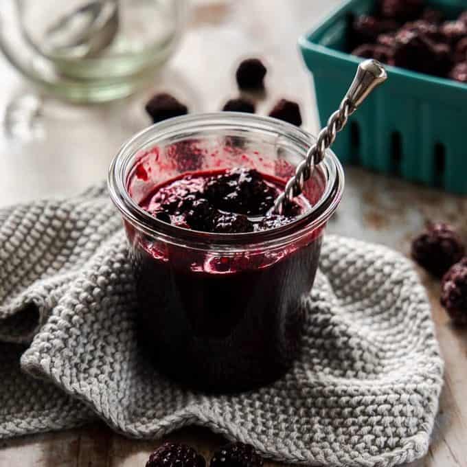 Front on view of a jar of blackberry compote with a spoon sticking out. It sits on a grey wash cloth.