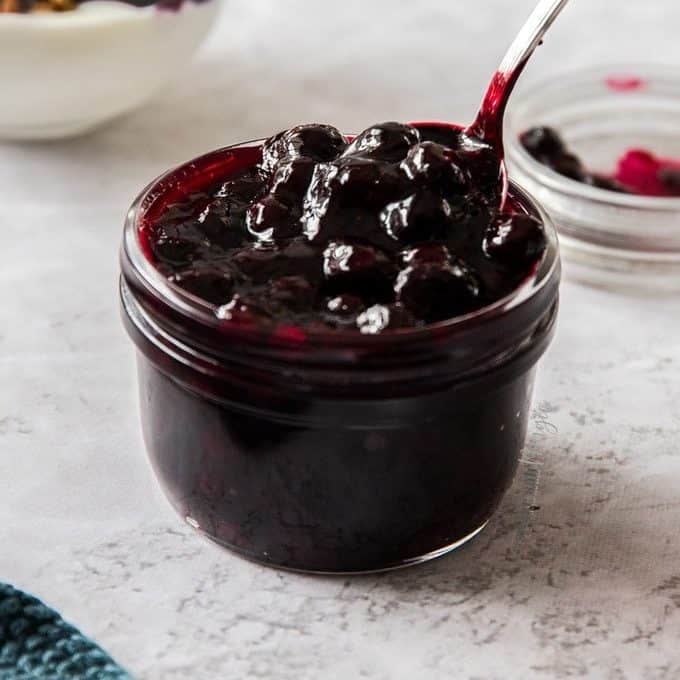 A small glass jar, filled to the brim with blueberry pie filling with a spoon digging in.
