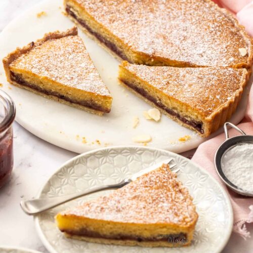 A bakewell tart cut into slices on a white marble cake platter.