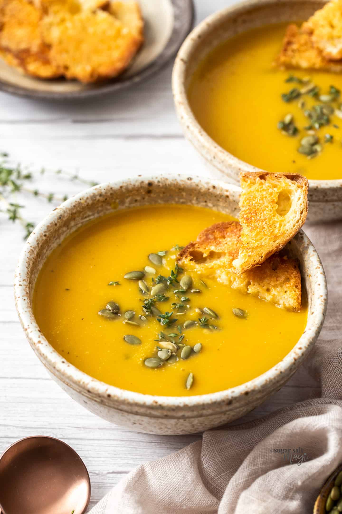 A bowl filled with butternut soup topped with crusty bread and pepitas. Another bowl behind it.