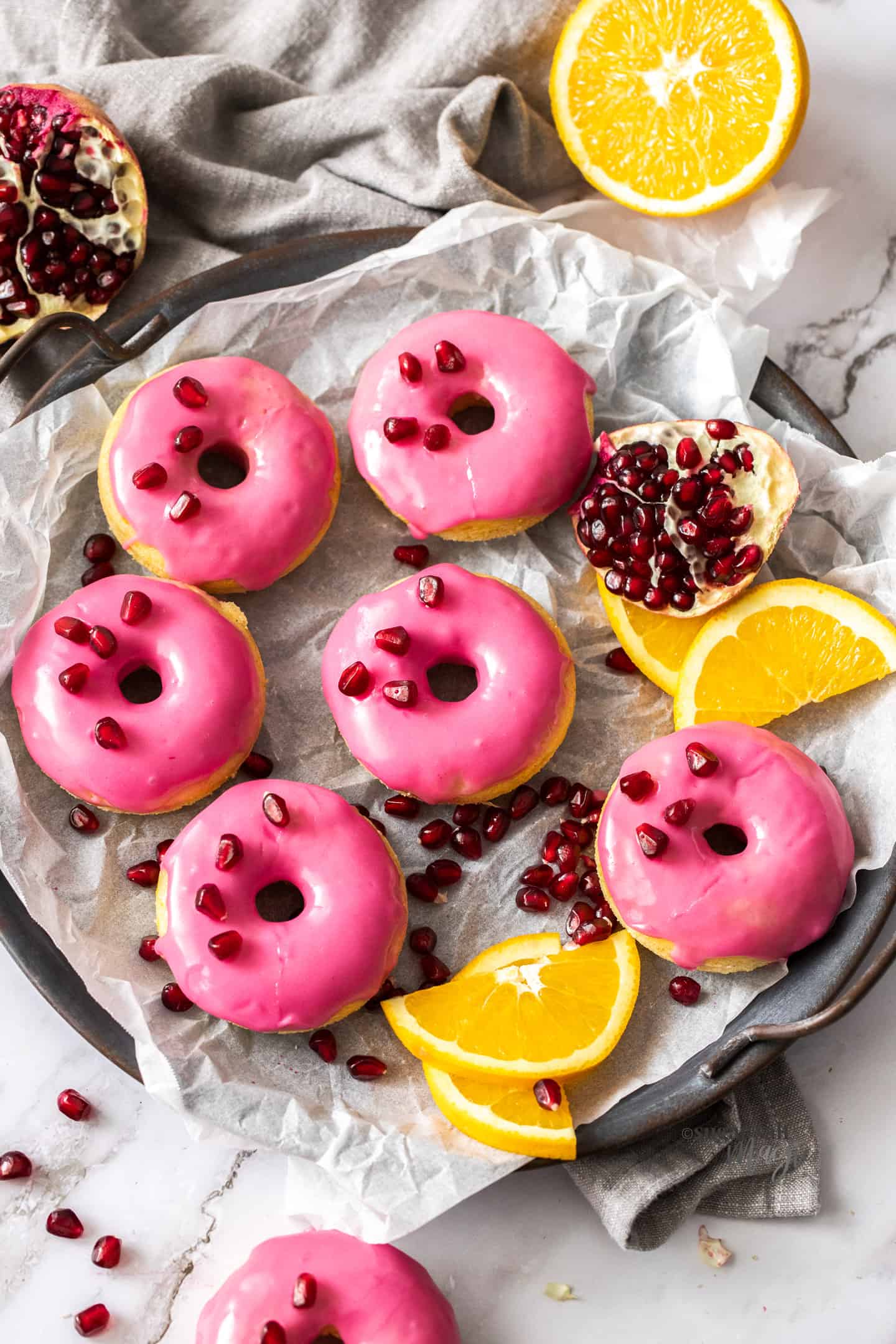 6 donuts with pink frosting, surrounded by orange slices and pomegranate seeds.
