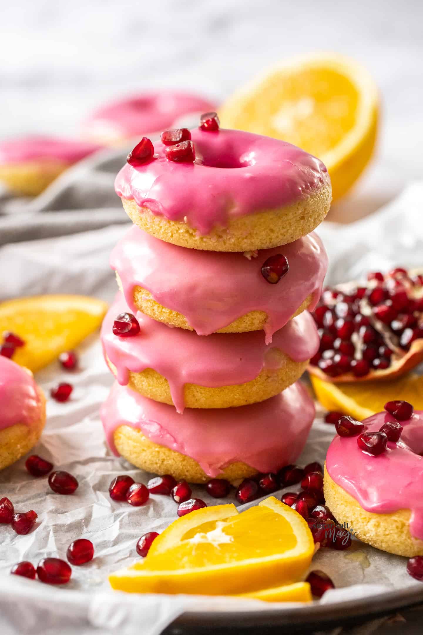 A stack of 4 donuts covered with pink icing. Pomegranate seeds are strewn around.