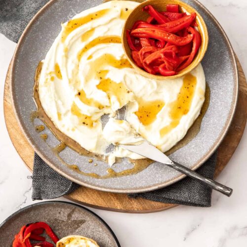 Goats cheese spread over a large grey plate with a small bowl of roasted peppers next to it.