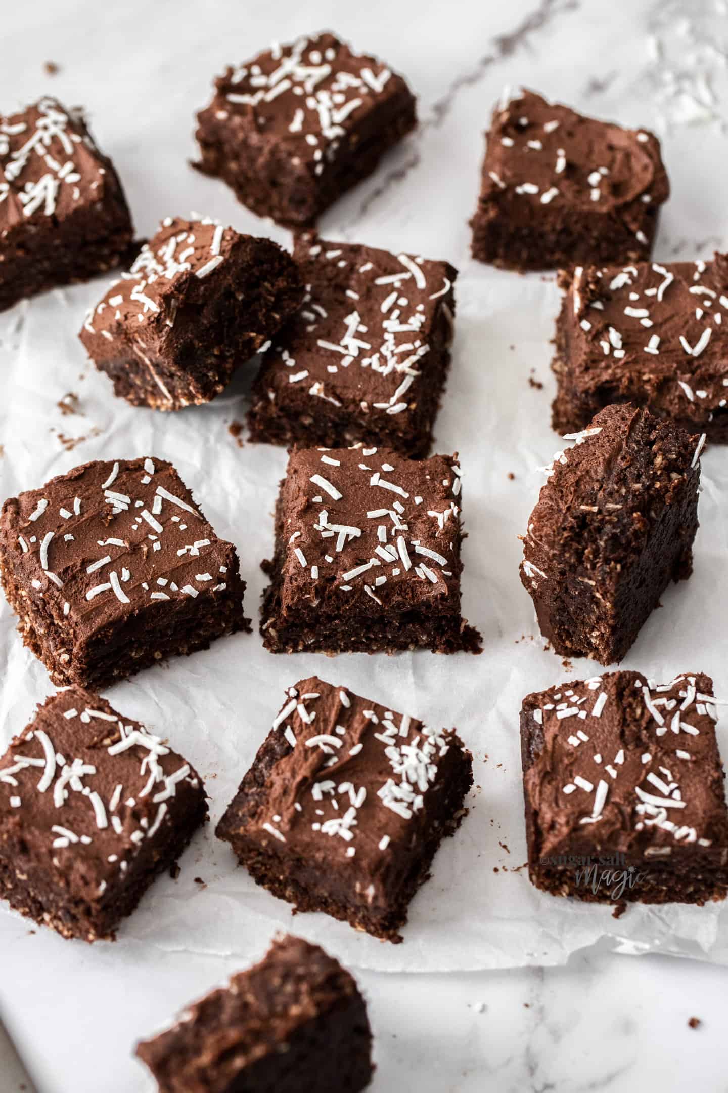 13 pieces of chocolate fudge slice topped with coconut on a sheet of baking paper.