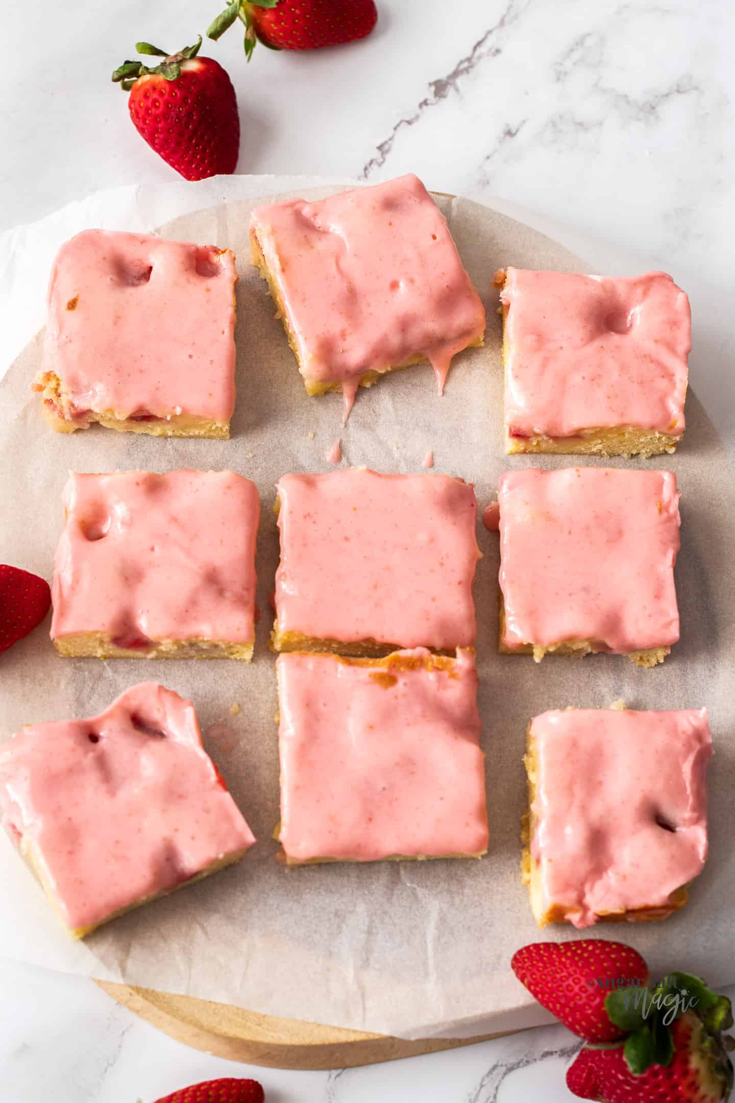 9 blondies with pink icing on a sheet of baking paper on a wooden board.