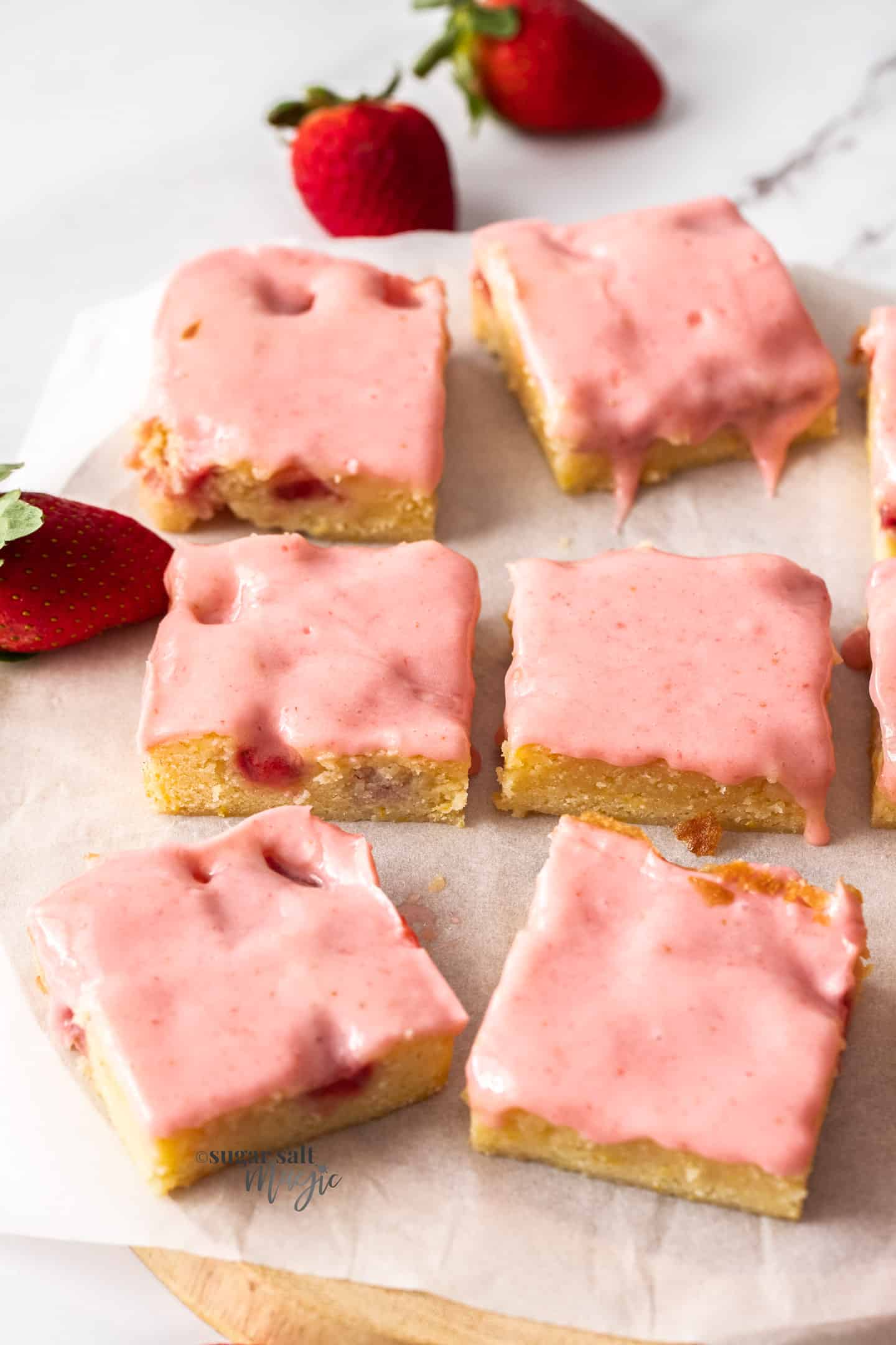 6 blondies with pink icing on a sheet of baking paper.