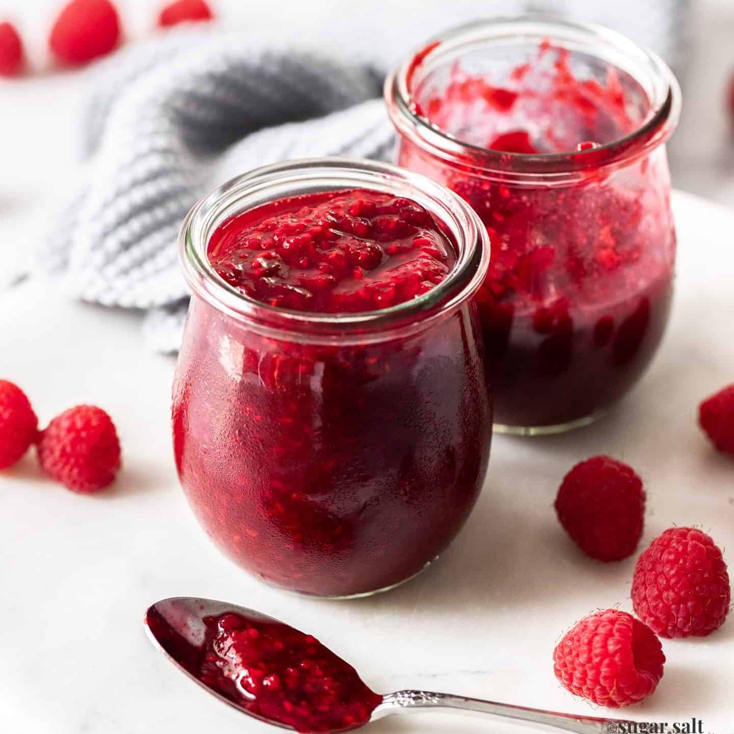 Two glass pots filled with raspberry compote and a spoonful in front.