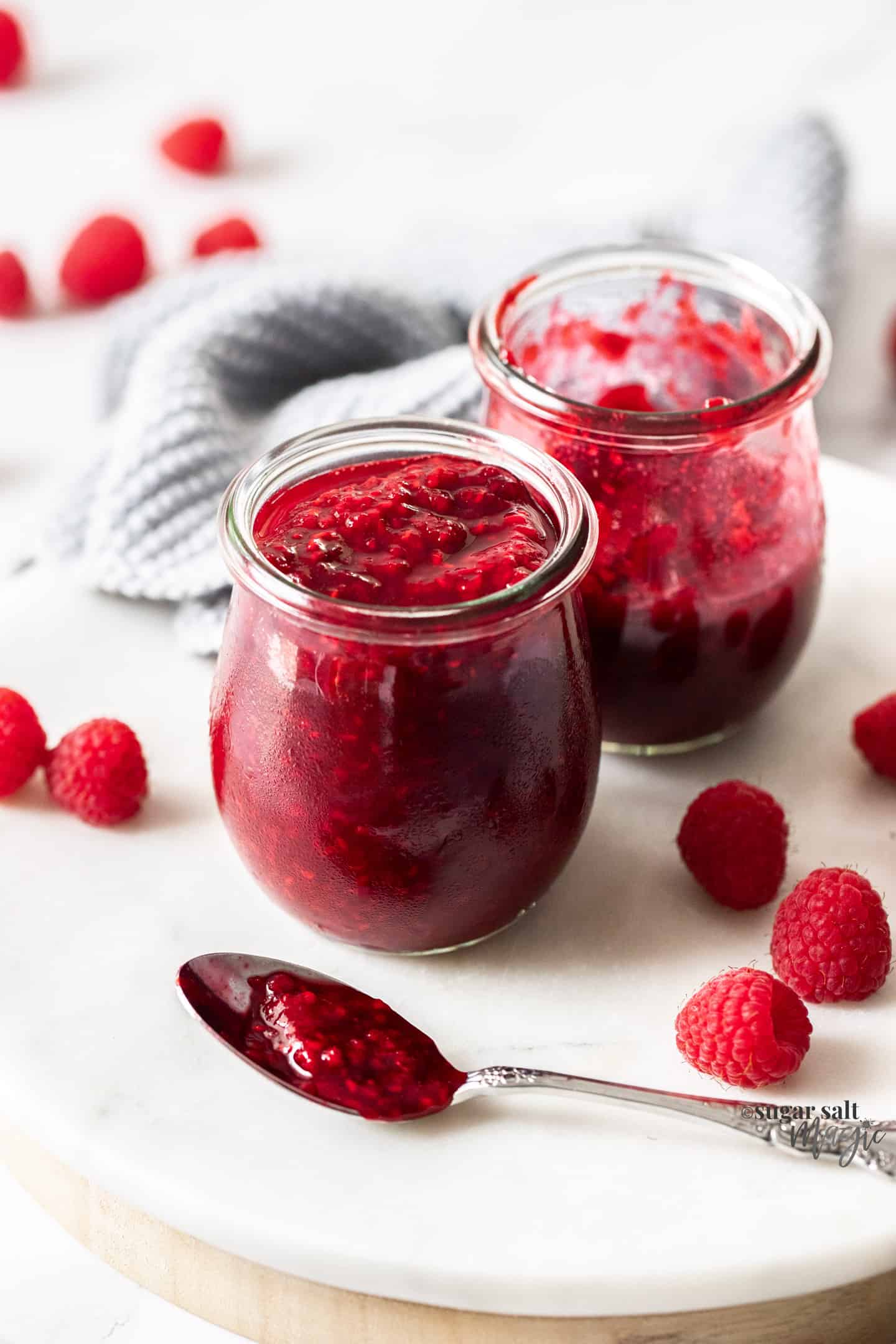 Two glass pots filled with raspberry compote and a spoonful in front.