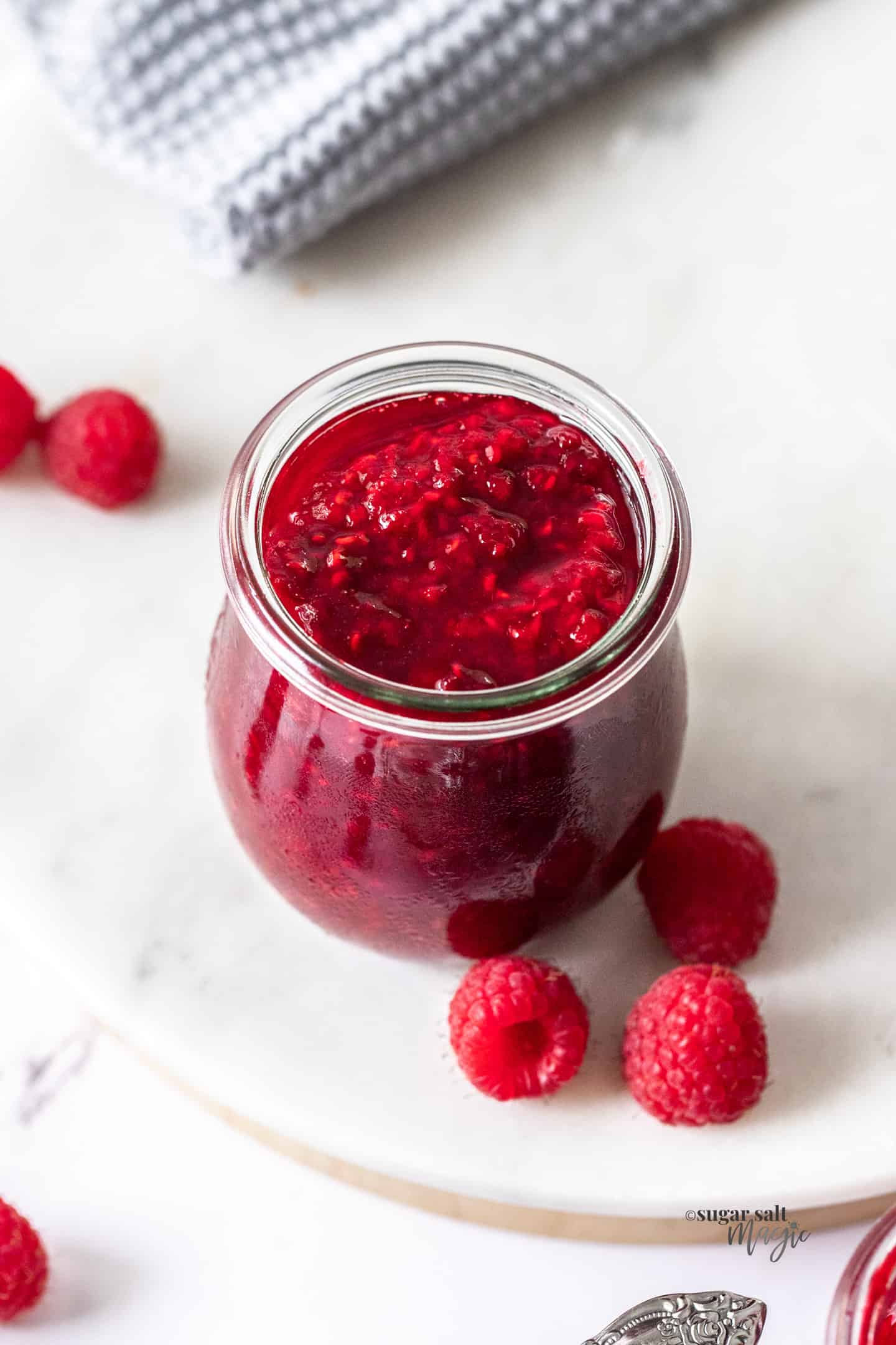 A small glass pot filled with raspberry compote. Fresh raspberries around.