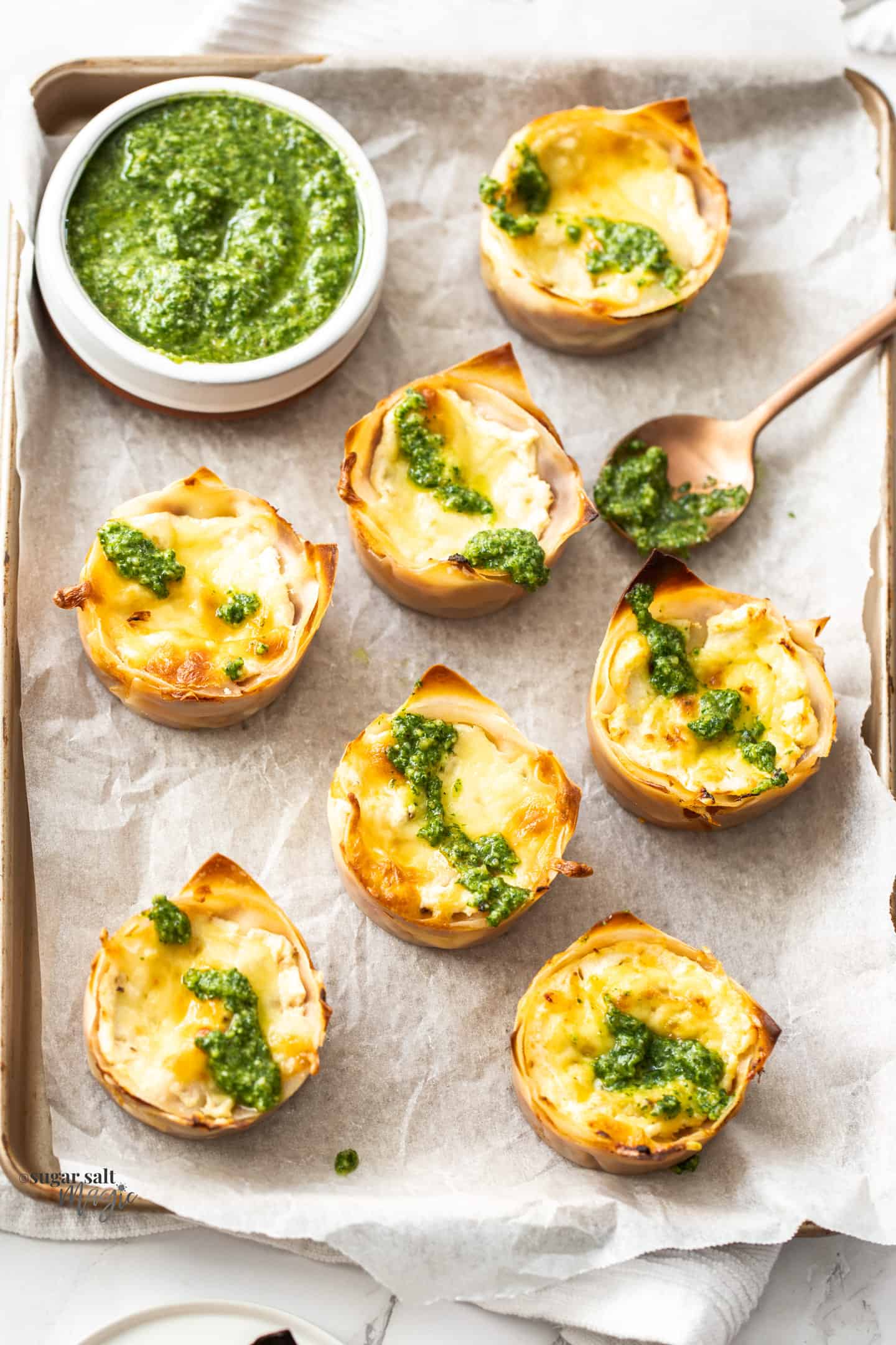 7 lasagna cupcakes sitting on a baking paper lined tray with a bowl of pesto.