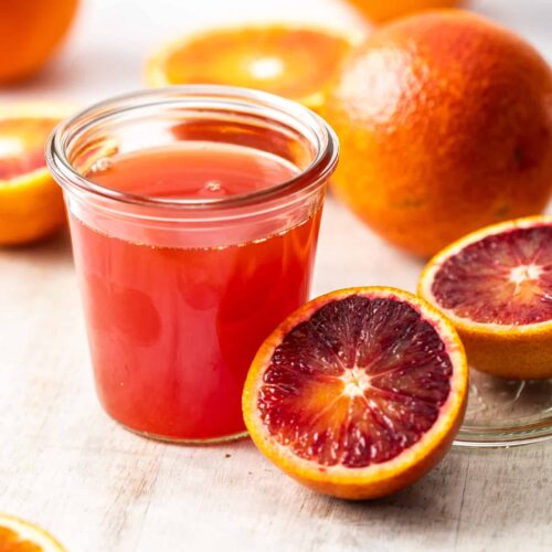 A preserving jar, filled with blood orange syrup surrounded by oranges.