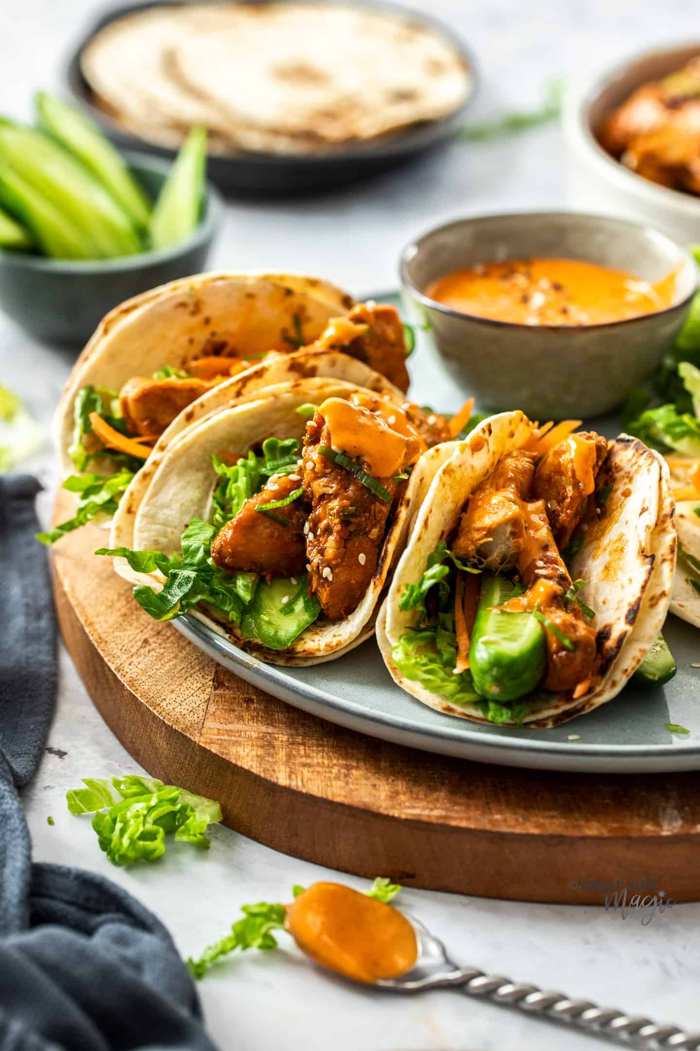 3 filled korean chicken tacos on a grey plate with a bowl of sauce behind.