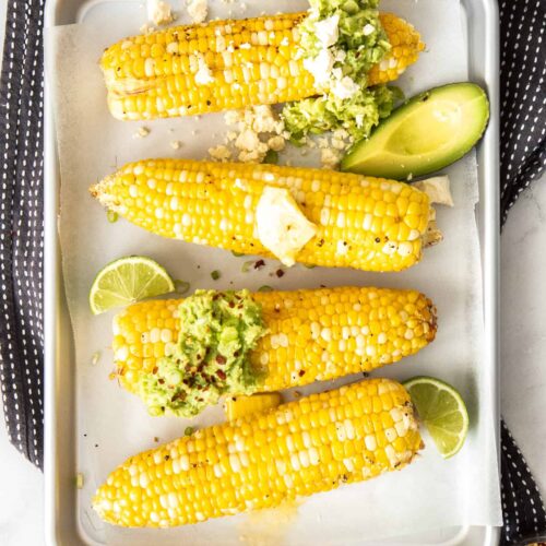 4 cobs of corn on a silver baking tray topped with butter and avocado.
