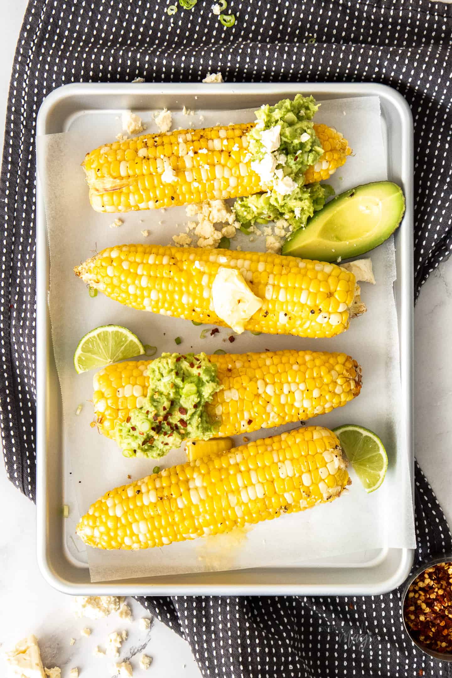 4 cobs of corn on a silver baking tray topped with butter and avocado.