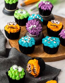 12 colourful cupcakes with candy eyes on a wooden platter.