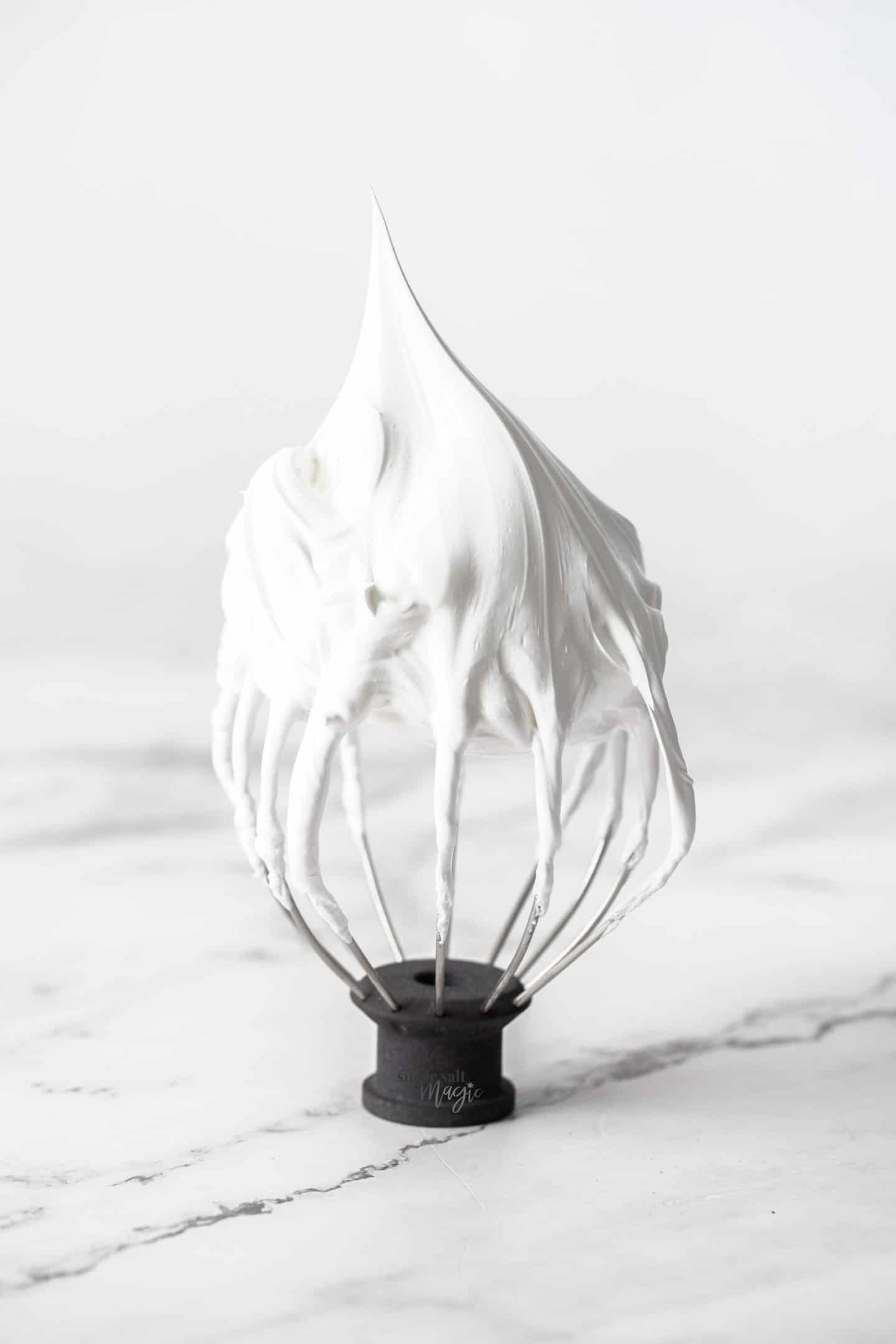 A whisk standing upright with a peak of meringue on top.