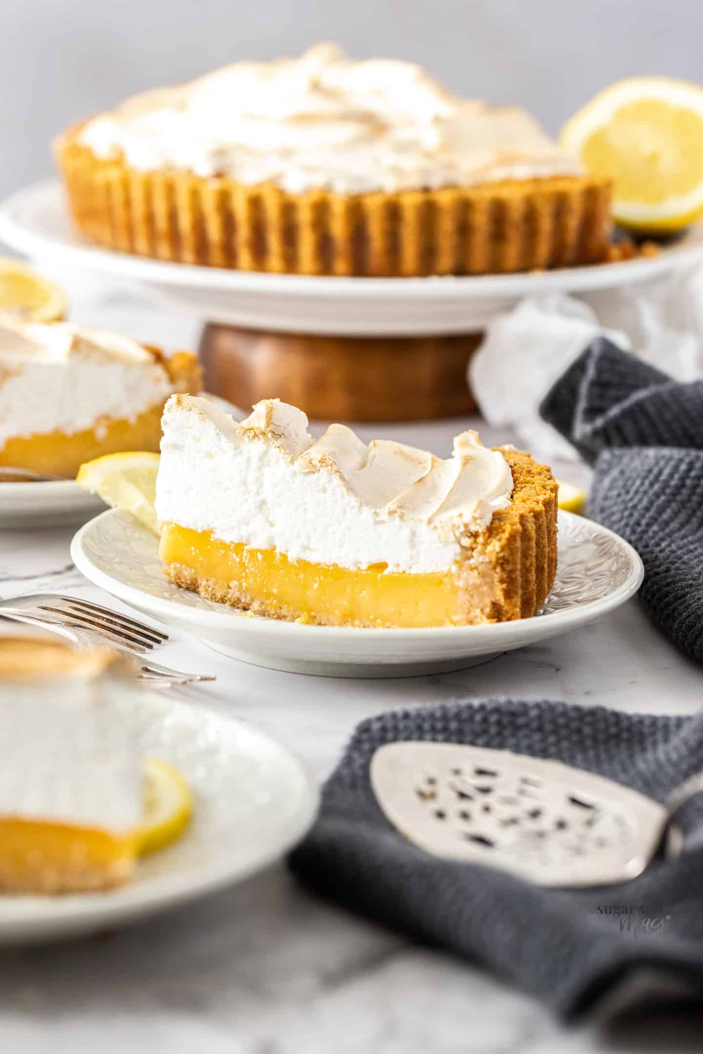 Slice of lemon meringue pie on dessert plates with the rest of it in the background.