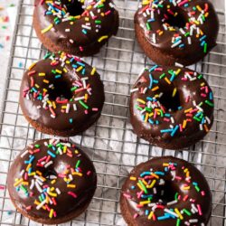 6 chocolate sprinkle donuts on a wire rack with a bowl of sprinkles behind.