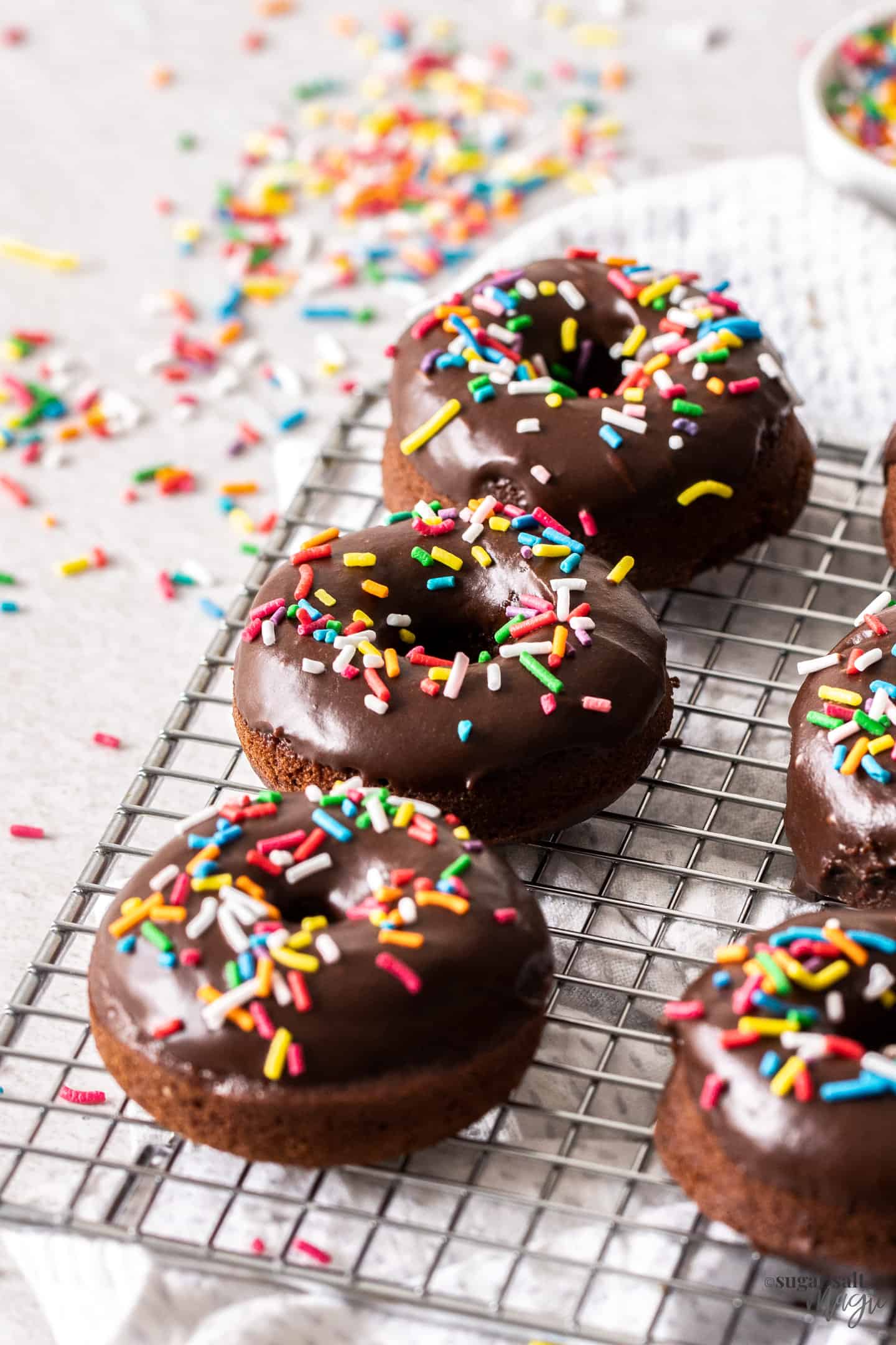 3 chocolate cake donuts covered in sprinkles on a wire rack.