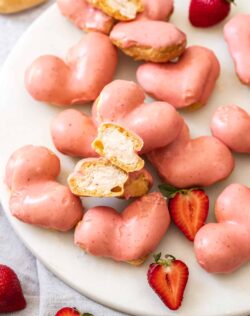 A pile of heart shaped cream puffs on a marble platter with one cut open.