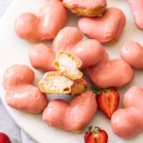 A pile of heart shaped cream puffs on a marble platter with one cut open.