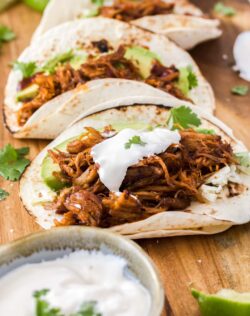 Pulled pork tacos on a wooden board with a bowl of sour cream.
