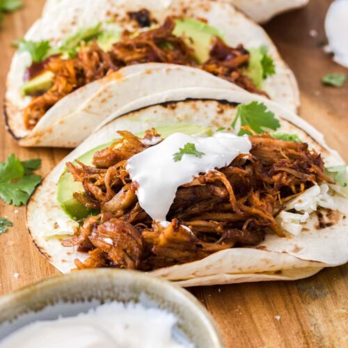 Pulled pork tacos on a wooden board with a bowl of sour cream.