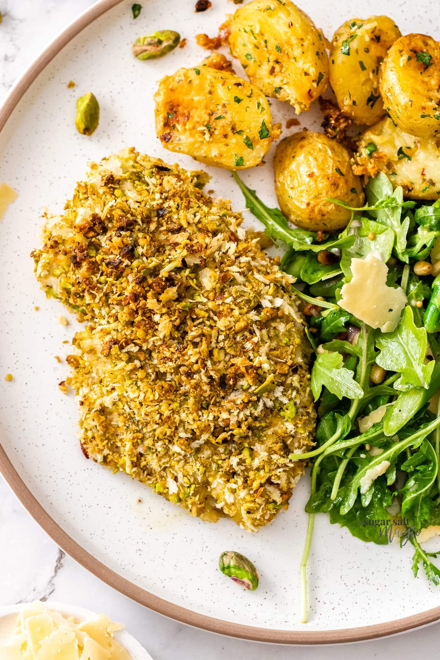 Closeup of pistachio crusted chicken breast next to potatoes and salad.