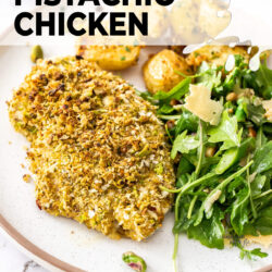 Close up of a baked chicken breast coated in pistachio crumbs next to a salad.