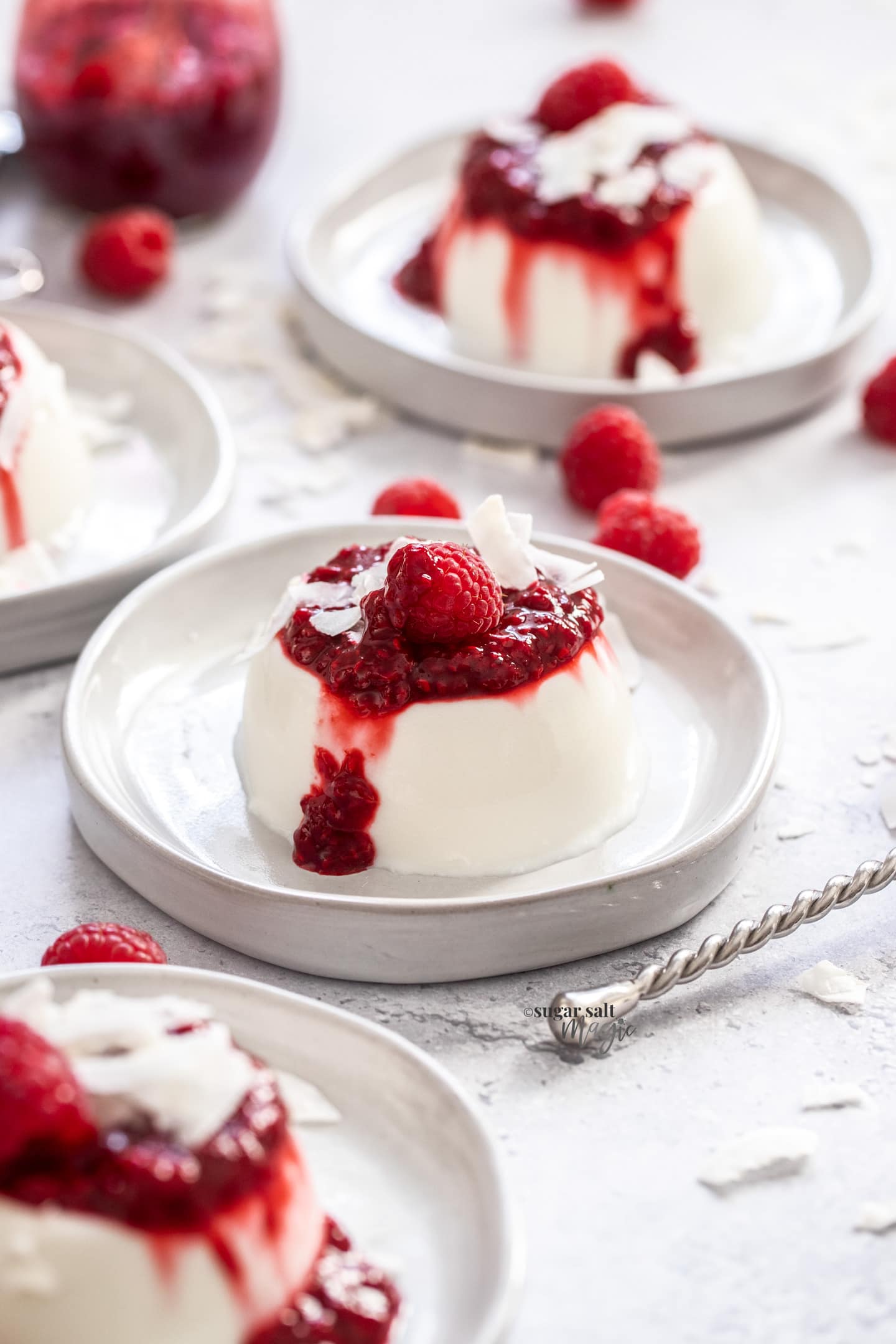 4 coconut panna cottas on dessert plates, topped with raspberry sauce.