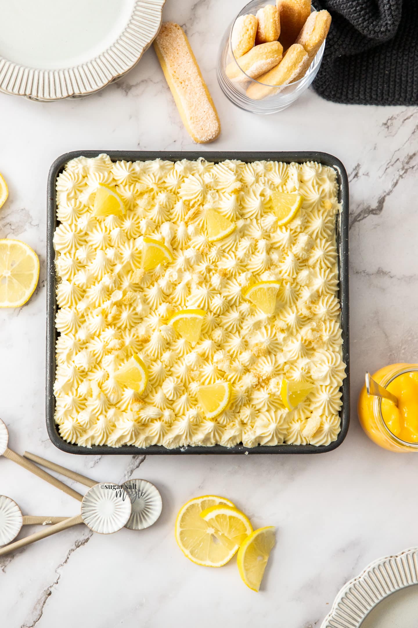 Top down view of lemon tiramisu in a pan surrounded by lemon slices and plates.