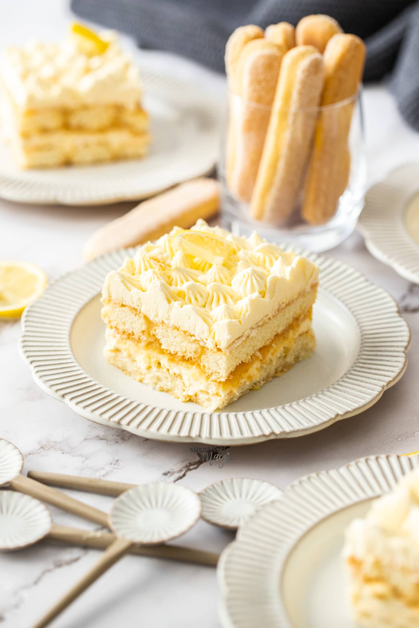 A square slice of lemon tiramisu on a small plate with a cup of lady fingers the background.