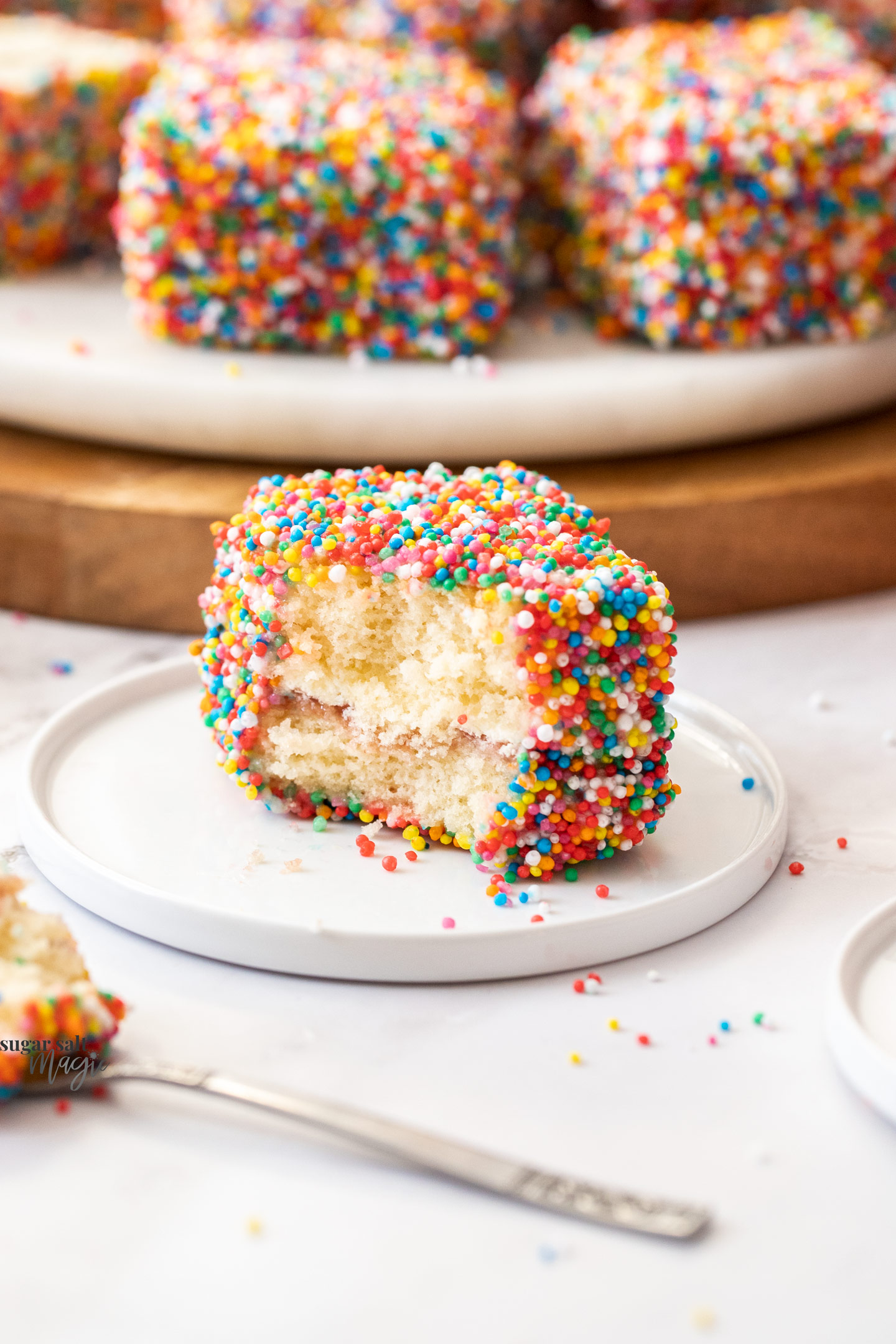 A fairy bread lamington on a small dessert plate with a chunk cut out.