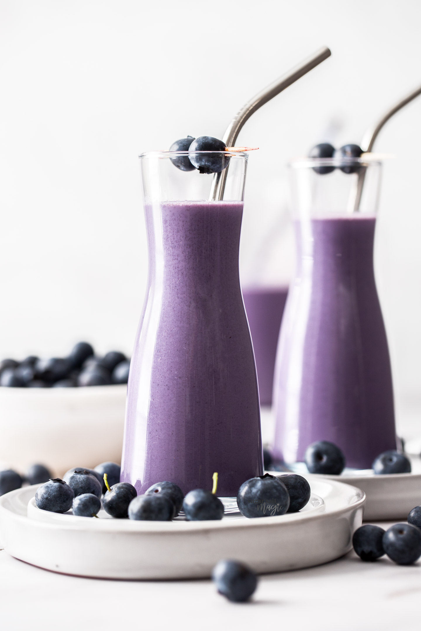 Two tall glasses filled with blueberry milk, surrounded by blueberries.