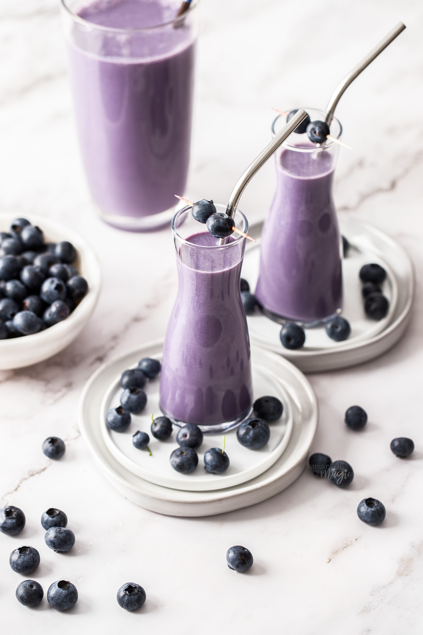Two tall glasses filled with blueberry milk with a jug in the background.