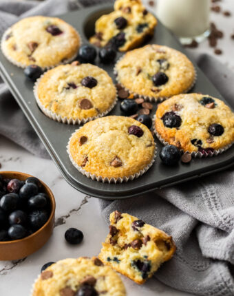 A muffin tin filled with baked blueberry muffins and surrounded by blueberries.
