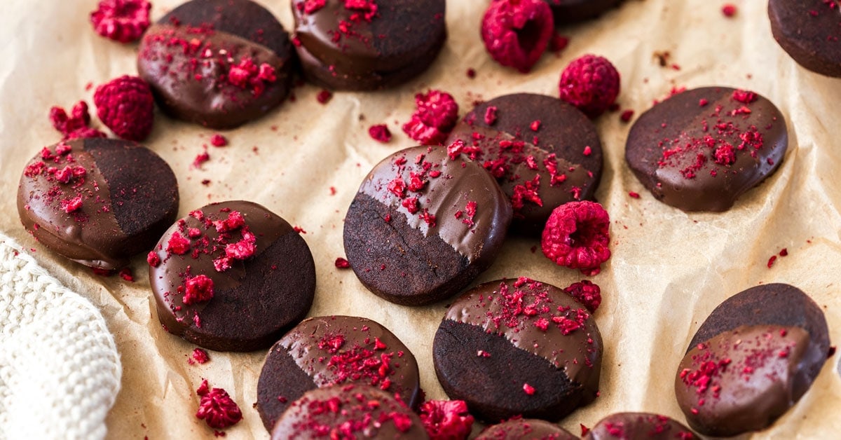 overhead: several dark chocolate shortbread cookies on a piece of brown parchment paper with freeze-dried raspberries on top