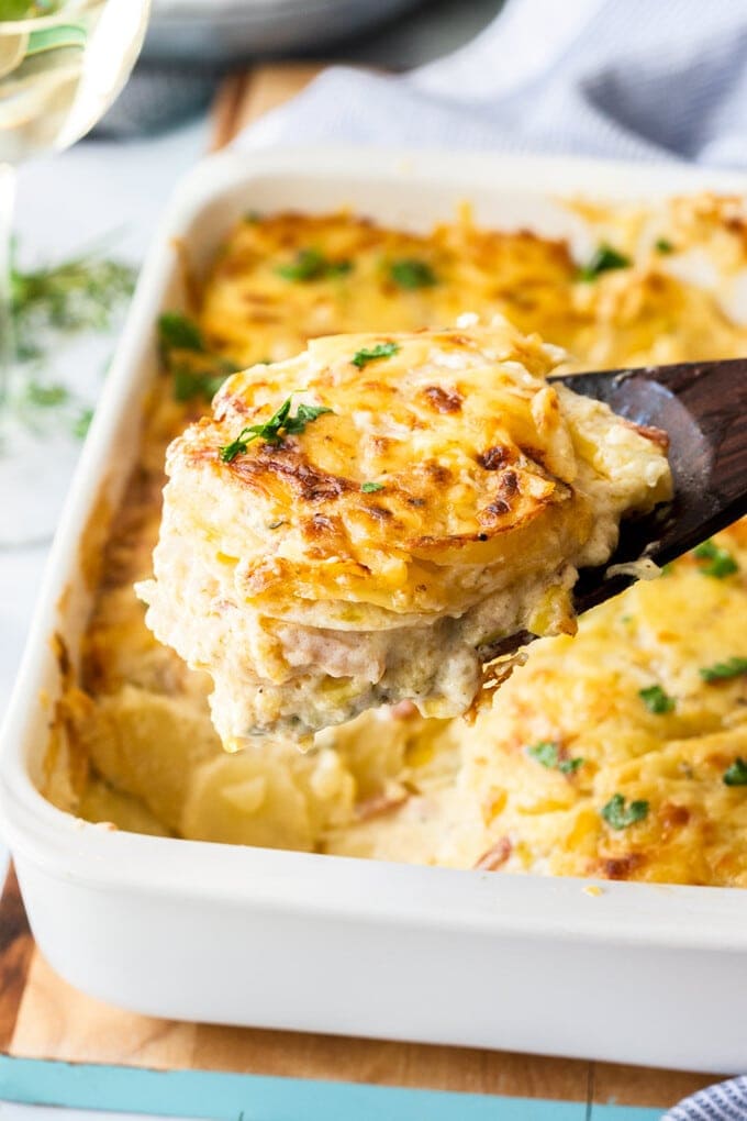 closeup: easy cheesy potatoes on a wooden spoon. A white casserole dish sits below it filled with scalloped potatoes.