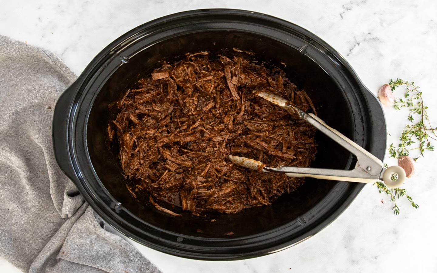 A slow cooker dish half full with shredded beef in gravy with a pair of tongs to the side.