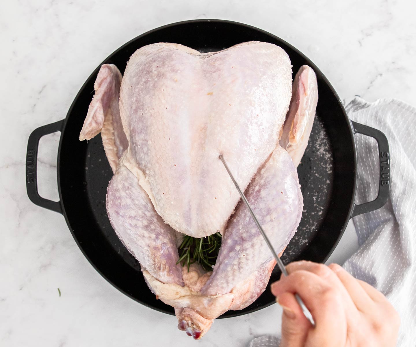 A skewer poking holes into the breast of a turkey in a large cast iron skillet.