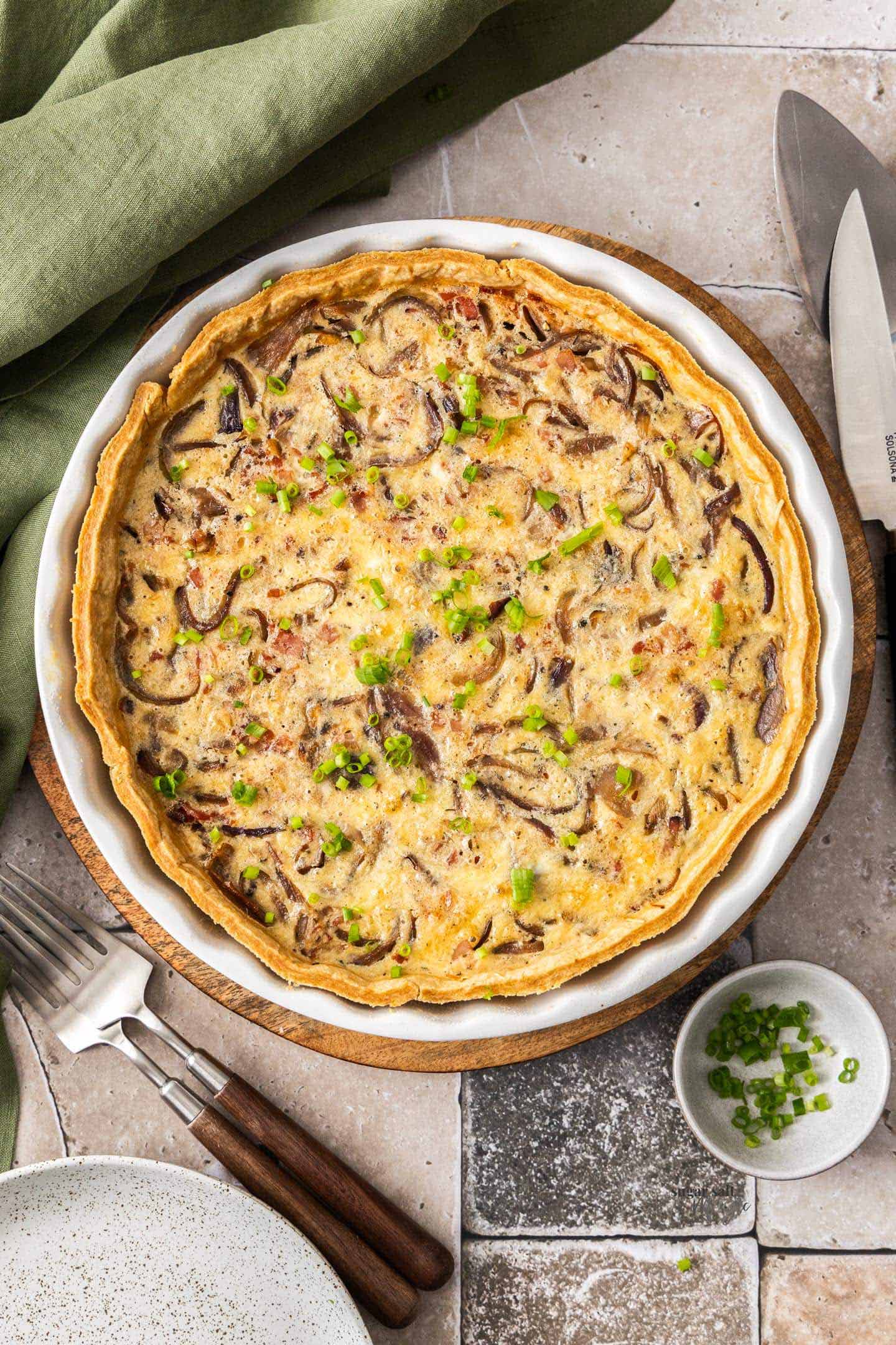 Golden flaky quiche with deeply caramelized brown onions and crispy bacon visible throughout the rich custard filling, melted Gruyère cheese on the surface with golden bubbly spots, fresh thyme sprigs as garnish, with a slice cut and lifted out showing the creamy interior.