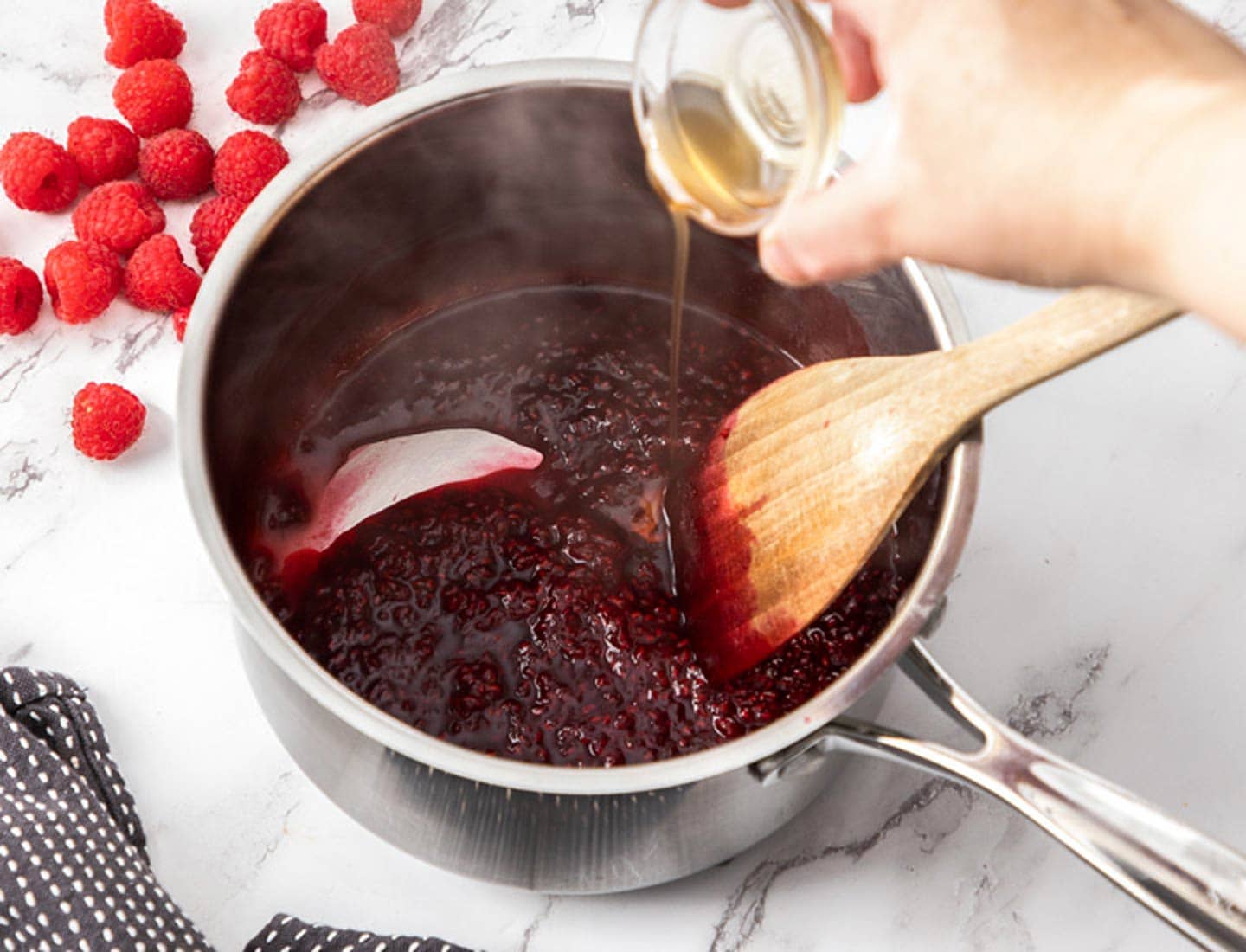 Coconut Raspberry Cake A hand pouring vanilla extract from a small bowl into a saucepan of raspberry compote.